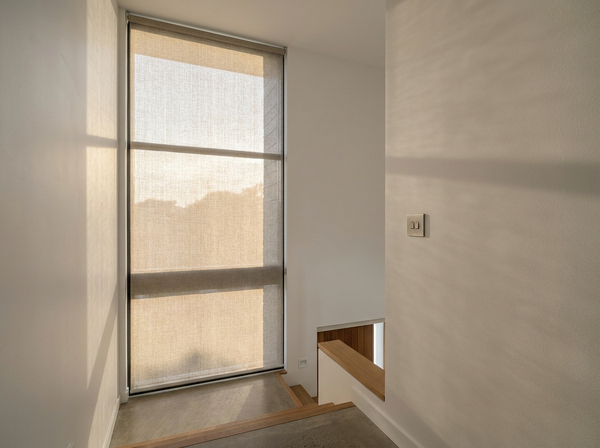 Motorised sunscreen roller blind lowered across a tall stairwell window with a wall-mounted control switch visible beside it
